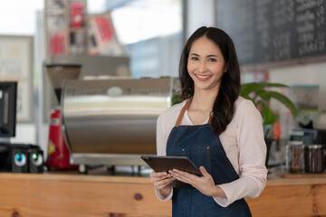 Asian female baristas wear aprons and hold tablet computer coffee menus at a counter bar with a smiling face, a cafe service concept.