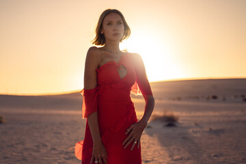 Romantic woman in a long red dress posing at a golden sunset light on the desert.
