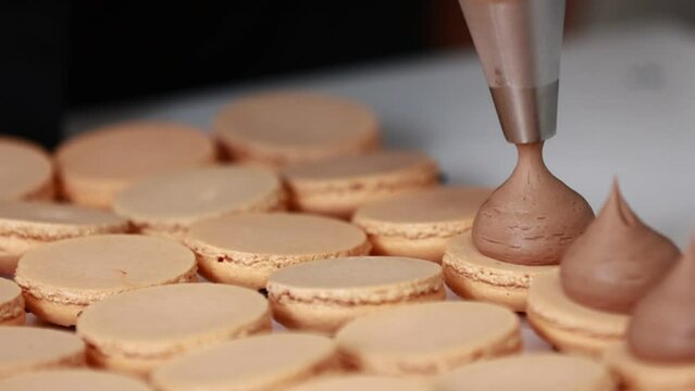 Making french desert macarons. Chef hands with confectionery bag add cream to the ready halves of brown chocolate macaroons at pastry shop. cooking, food and baking concept