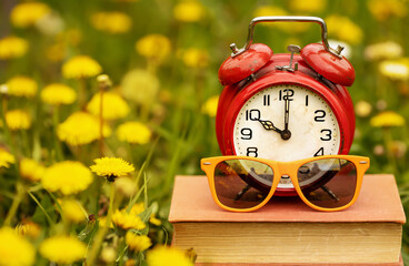 Alarm clock and sunglusses on a book with yellow flowers in the grass. End of school, summer break time.