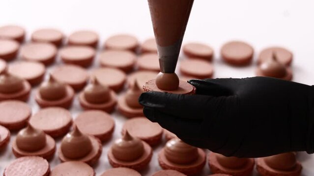 Making french desert macarons. Chef hands with confectionery bag add cream to the ready halves of brown chocolate macaroons at pastry shop. cooking, food and baking concept