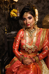 Magnificent young Indian bride in luxurious bridal costume with makeup and heavy jewellery sitting in a chair with classic vintage interior in studio lighting. Wedding Fashion and Lifestyle.