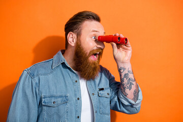 Photo of positive excited guy dressed denim jacket looking binoculars empty space isolated orange color background