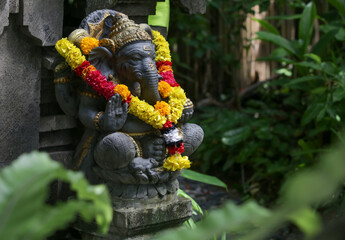 Traditional Ganesha statue with flower wreath in Balinese garden. Lord Ganesha is believed to bring...