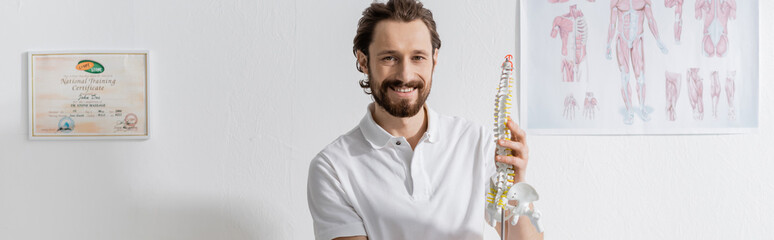 Obraz premium cheerful chiropractor with spine model smiling at camera in consulting room of rehab center, banner.