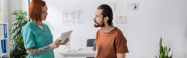 tattooed physiotherapist with digital tablet talking to bearded man in consulting room, banner.