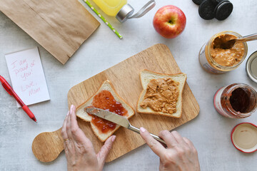 Mom making PB&J sandwich while packing lunch for child. Flat lay food apple, note of encouragement