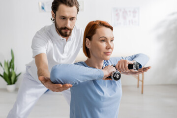 rehabilitologist assisting redhead woman working out with dumbbells in recovery center.