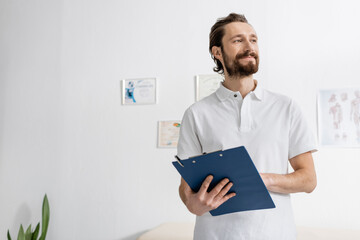 pleased bearded osteopath holding clipboard and looking away in rehabilitation center.