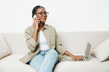 African American woman business freelancer working sitting on the couch at home in a laptop and phone, business calls and correspondence sadness and anger, home clothes and eyeglasses, light interior