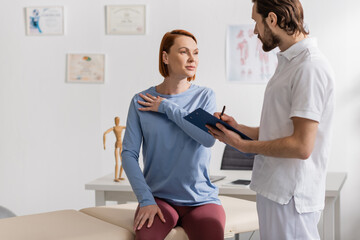 redhead woman touching painful shoulder near physiotherapist writing prescription on clipboard in consulting room.