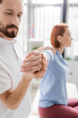 Fototapeta premium bearded physiotherapist stretching painful arm of blurred woman during diagnostics in consulting room.