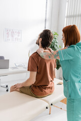 redhead osteopath touching painful back of man sitting on massage table in consulting room. - obrazy, fototapety, plakaty