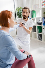 bearded physiotherapist helping redhead woman working out with dumbbells in recovery clinic.