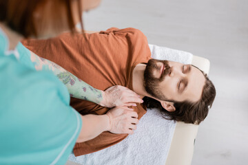 high angle view of bearded man with closed eyes near blurred physiotherapist doing shoulder massage in consulting room.
