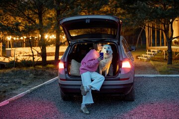 Portrait of a young woman with her cute white dog sitting together at car trunk, traveling on...