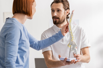 bearded osteopath holding spine model and talking to redhead woman in consulting room.