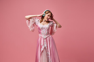 Shot of adorable blond princess, queen wearing fancy dress and listening music over pink studio background. Modern princess