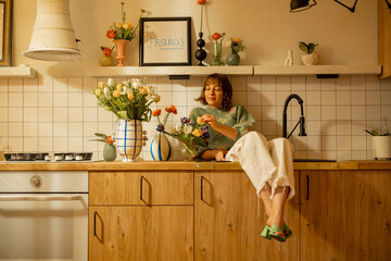 Cute and relaxed woman sits on kitchen sink in modern kitchen interior decorated with fresh...