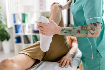 cropped view of tattooed chiropractor flexing leg of patient during treatment in rehabilitation center.