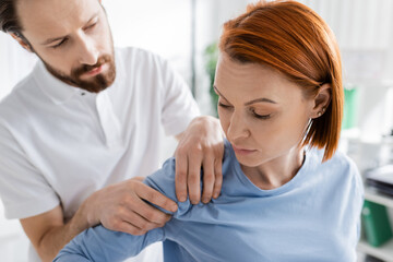 physiotherapist touching injured shoulder of redhead woman during diagnostics in consulting room.