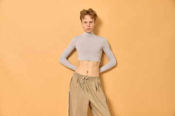Cocky caucasian gay man with short curly red hair wearing trendy beige crop top and pants standing with bare belly near yellow wall with hands behind his back and looking at camera with confidence.