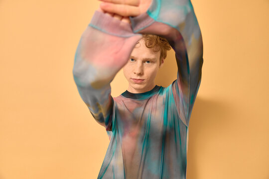 Androgynous queer young man with red curly hairstyle wearing gradient vivid translucent bodysuit, stretching his body outstretching his arms while dancing vogue on yellow backdrop in studio. Lgbtq.