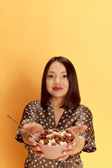 Portrait of young asian girl in comfortable homewear, holding bowl with cereal against yellow studio background. Morning, breakfast. Concept of emotions, lifestyle, youth, happiness. Calm face