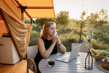 Smiling Woman freelancer using a laptop on a cozy glamping tent in a sunny day. Luxury camping tent for outdoor summer holiday and vacation. Lifestyle concept