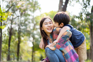 Fototapeta premium Asian woman and boy playing and walking around the park together with beautiful bokeh and lighting background copyspace. Little cute boy hogging his mother from back and kissing.