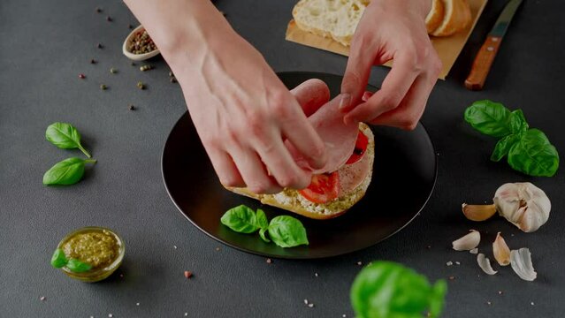 Men's hands prepare Chiabatta Sandwich with ham, basil, pesto sauce, tomatoes on a black background. Food background