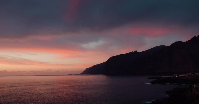 Dramatic cloudscape at sunset with reflection on ocean surface of pink clouds sky. Scenic viewpoint on sea water waves. Steep cliff bluff, Tenerife coast, Spain.