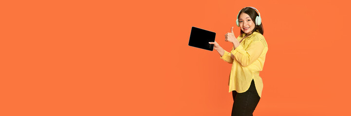 Portrait of smiling, young, positive, asian girl in headphones, holding tablet against orange studio background. Concept of emotions, lifestyle, youth, sales. Banner. Copy space for ad