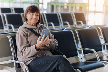 Smiling adult traveler woman in eyeglasses awaits boarding a flight for departure while uses smartphone with free wi fi at airport terminal. Middle aged female independently traveling abroad.