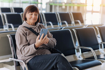 Smiling adult traveler woman in eyeglasses awaits boarding a flight for departure while uses...