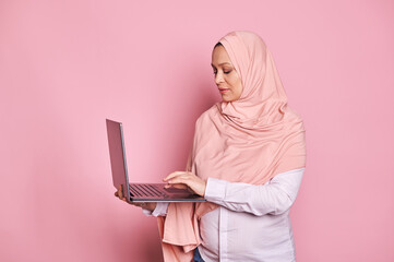 Overworked multitasking Arab Muslim pregnant woman, manager, business lady, entrepreneur, dressed in pink hijab, typing text on laptop keyboard, isolated over pink background. Pregnancy and career