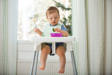 Cute baby eating first solid food, infant sitting in high chair. Child tasting vegetables at the table, discovering new food. Cozy kitchen interior. Healthy food concept. 