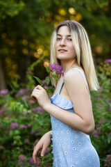 Beautiful blonde girl looking at camera, dressed in summer slim dress, holding in hands a flower, park background.