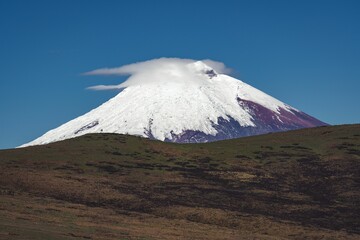 Fototapeta premium Layer of white fluffy clouds in front of snowy Cotopaxi volcano on blue sky background in Ecuador