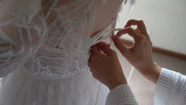 The bride wears a wedding dress. A young girl in a white lace luxury dress in the morning. The bridesmaid or mother tying lace on the back of the corset.