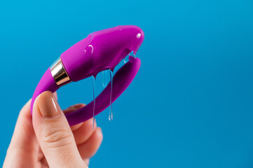 A woman holds a curved sex toy in lubrication on a blue background. Vaginally clitoral vibrator. - obrazy, fototapety, plakaty