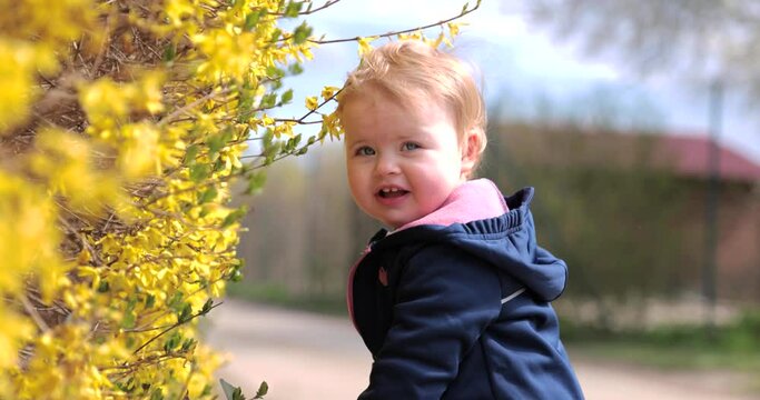Beautiful baby girl playing with yellow flowers in nice spring sunny day outdoor. Blonde hair blue eyes todler. Slow motion shot 4K 120fps, high quality. Bouncing tree branches. Discovering new things