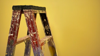 Old used construction painter ladder with painted splatter standing in front of the yellow wall under warm white lighting © Darr.di