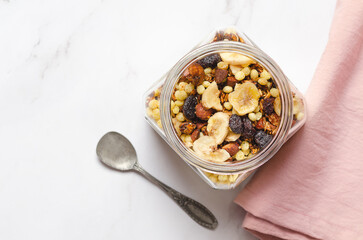 Homemade granola with nuts, dried banana slices and dried cranberries in a glass jar with a pink napkin.