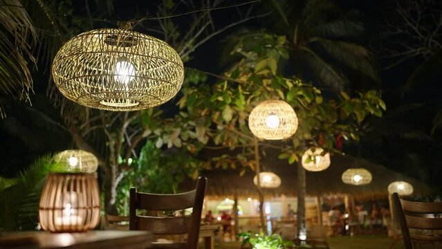 Wicker straw lamps hanging from trees as decoration against tropical background at night. Old-fashioned wooden bulbs sway in wind lighting outdoor cafe. Boho wedding decorations at exotic setting