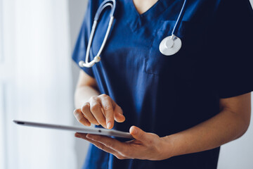 Doctor woman using tablet computer while standing near panorama window in clinic, close up. Physician or surgeon at work. Medicine concept