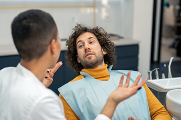 Young man with curly hair communicating carefully listening and looking at his dentist during appointment consultations for dental treatment and future procedures at dentist's office.