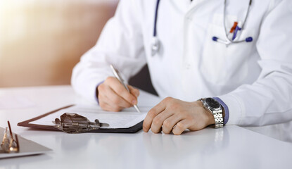 Unknown male doctor sitting and working with clipboard of medication history record in a darkened clinic, glare of light on the background, close-up of hands