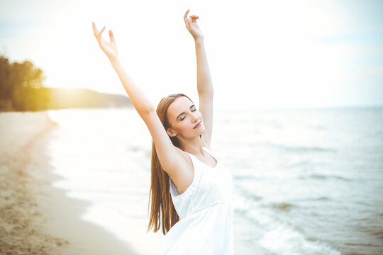Happy smiling woman in free happiness bliss on ocean beach standing with raising hands. Portrait of a multicultural female model in white summer dress enjoying nature.