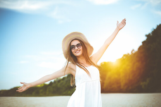Happy smiling woman in free happiness bliss on ocean beach standing with a hat, sunglasses, and open hands. Portrait of a multicultural female model in white summer dress enjoying nature